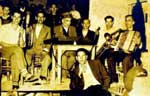 Γαμήλιο γλέντι στο παλιό καφενείο "του Καμαρού" στην Αγιάσο το 1954. Παίζουν οι Αγιασώτες μουσικοί, Μιχάλης Μουτζουρέλλης ("Λαγός") - κλαρίνο, Ραφαήλ Σουσαμλής - ευφώνιο, Στρατής Ρόδανος - σαντούρι, Αχιλλέας Σουσαμλής - βιολί, Στρατής Αλτιπαρμάκης ("Ρογίδι") - τρομπόνι και Στρατής Παπάνης - ακορντεόν. Αρχείο Μιχ. Μουτζουρέλλη.